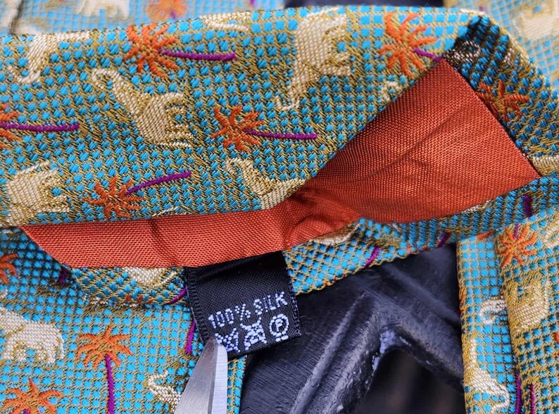 May include: Close-up of a silk tie with a teal background featuring a pattern of elephants and palm trees in gold and orange. The tie has a red lining and a label that reads "100% Silk".