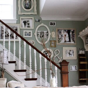 A gallery-style display of white-framed artwork on a green wall next to a set of stairs. The art includes framed prints and photos in various shapes and sizes, mirrors, and other miscellaneous pieces placed in an appealing arrangement.