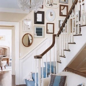 Example of a foyer with a gallery-style display of numerous mirrors of various shapes and sizes in white, gold or black frames hung on a light beige wall next to a set of stairs.