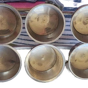 May include: Six small, round, metal containers with a shiny, silver finish. The containers are arranged in a semi-circular pattern, with their openings facing upwards. The interior of each container is visible, showing a smooth, reflective surface.