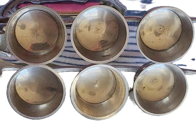 May include: Six small, round, metal containers with a shiny, silver finish. The containers are arranged in a semi-circular pattern, with their openings facing upwards. The interior of each container is visible, showing a smooth, reflective surface.