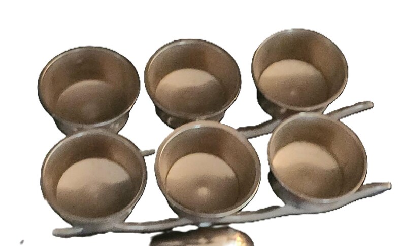 May include: A silver-toned metal serving piece with six small, round bowls. The bowls are arranged in two rows of three, connected by a branching stem. The interior of each bowl appears to be a light beige color.
