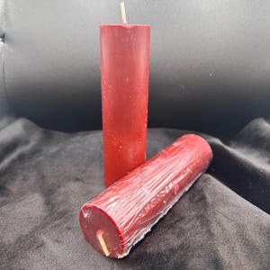 May include: Two red pillar candles with a wick in the centre of each. The candles are wrapped in clear plastic.