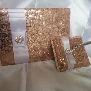 May include: A rose gold sequined guest book with a white satin ribbon and a rhinestone embellishment. A matching rose gold sequined pen holder with a white satin ribbon and a rhinestone embellishment.
