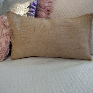 Puede incluir: Una almohada de arpillera marr&oacute;n con una superficie texturizada. La almohada est&aacute; sobre una colcha blanca acolchada.