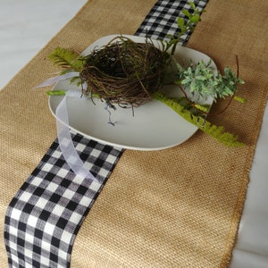 May include: A burlap table runner with a black and white checked ribbon down the centre. A white plate with a twig nest and green sprigs is on top of the runner.
