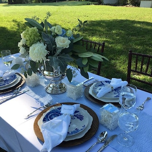 May include: A table setting with white and blue china plates, white napkins, and a centerpiece of white and green flowers in a silver vase. The table is set on a blue and white checkered tablecloth. The table is outdoors on a grassy lawn.