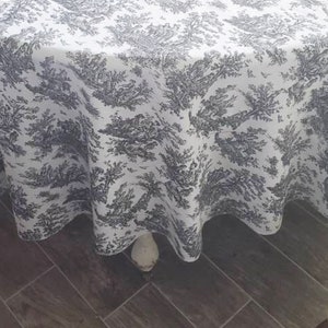 May include: A white tablecloth with a black and white toile pattern. The tablecloth is round and has a scalloped edge. The tablecloth is draped over a table and the edge of the tablecloth is hanging down.