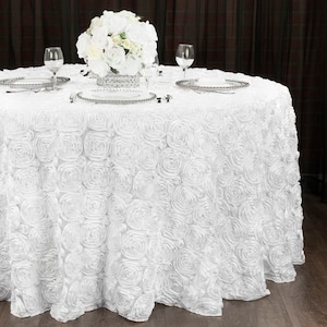 May include: White round tablecloth with a rosette design. The tablecloth is made of satin and has a ruffled edge. A white flower arrangement is on the table.