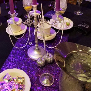 May include: A silver candelabra with three purple candles, a silver serving dish, and two white teacups on a purple sequined tablecloth. There are also silver pumpkins and a silver string of pearls on the table.