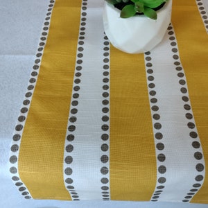 May include: A yellow and white striped table runner with a repeating pattern of small, dark brown dots. The runner is made of a textured fabric and is decorated with a small white pot with a green plant.