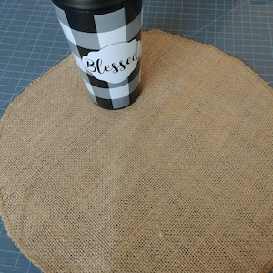 May include: A black and white buffalo check tumbler with the word "Blessed" in a white oval. The tumbler sits on a round, tan burlap placemat. The placemat has a woven texture.