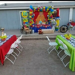 May include: A red and green table setting with a black and white cow print runner. The table is set with white folding chairs. The background features a Toy Story themed party decoration with balloons, a red curtain, and a hay bale.