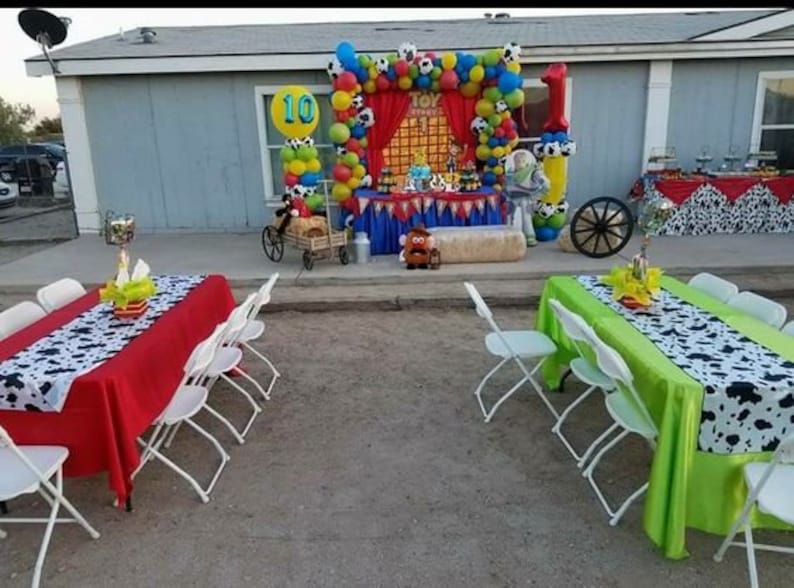 May include: A red and green table setting with a black and white cow print runner. The table is set with white folding chairs. The background features a Toy Story themed party decoration with balloons, a red curtain, and a hay bale.