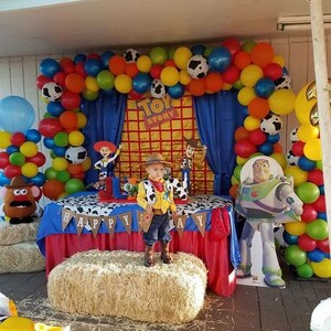 May include: A Toy Story themed birthday party with a blue and red table cloth, a hay bale, and a balloon arch. The arch is made of red, yellow, blue, green, and white balloons. There is a cardboard cutout of Buzz Lightyear and a child dressed as Woody.