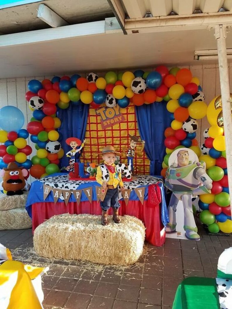 May include: A Toy Story themed birthday party with a blue and red table cloth, a hay bale, and a balloon arch. The arch is made of red, yellow, blue, green, and white balloons. There is a cardboard cutout of Buzz Lightyear and a child dressed as Woody.