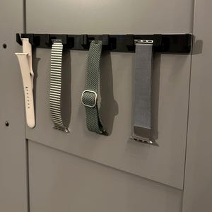 May include: A black watch band organizer displaying four watch bands. The bands include a light pink silicone band, a silver metal link band, a green fabric band with a silver buckle, and a silver mesh band.