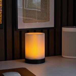 Op de afbeelding: Een cilindrische tafellamp met een geplooide witte kap en een zwarte voet. De lamp straalt een warme, gele gloed uit. De lamp staat op een wit bureau met een bruine leren muismat en een witte computermuis.