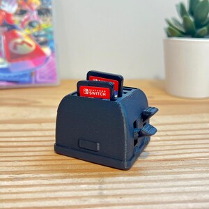 May include: A black 3D-printed toaster-shaped game cartridge holder with two red Nintendo Switch game cartridges inserted. The holder has two lever-like protrusions and is sitting on a wooden surface. A game case and a small potted plant are in the background.