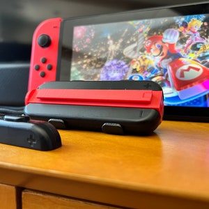 K&ouml;nnte beinhalten: Eine Nintendo Switch-Konsole mit roten Joy-Con-Controllern und einem schwarzen Griff. Der Bildschirm zeigt ein farbenfrohes Rennspiel mit Mario und Prinzessin Peach. Die Konsole steht auf einer Holzoberfl&auml;che.