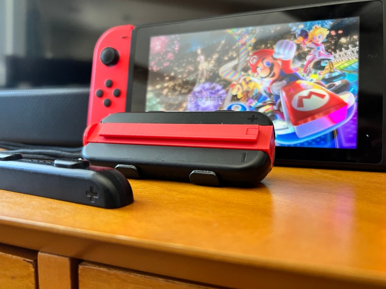 K&ouml;nnte beinhalten: Eine Nintendo Switch-Konsole mit roten Joy-Con-Controllern und einem schwarzen Griff. Der Bildschirm zeigt ein farbenfrohes Rennspiel mit Mario und Prinzessin Peach. Die Konsole steht auf einer Holzoberfl&auml;che.