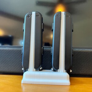 K&ouml;nnte beinhalten: Ein wei&szlig;er, 3D-gedruckter St&auml;nder, der zwei schwarz-wei&szlig;e Gamecontroller h&auml;lt. Die Controller haben ein Plus- und Minus-Symbol oben. Der St&auml;nder steht auf einer Holzoberfl&auml;che.