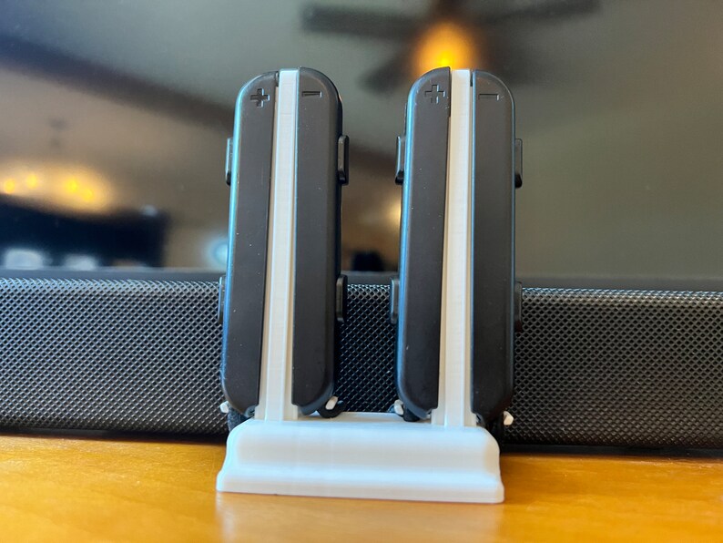 K&ouml;nnte beinhalten: Ein wei&szlig;er, 3D-gedruckter St&auml;nder, der zwei schwarz-wei&szlig;e Gamecontroller h&auml;lt. Die Controller haben ein Plus- und Minus-Symbol oben. Der St&auml;nder steht auf einer Holzoberfl&auml;che.