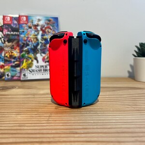 May include: A pair of red and blue Nintendo Switch Joy-Con controllers stand upright. Behind them are three game cases, with a small potted succulent plant to the right. The controllers are on a wooden surface.