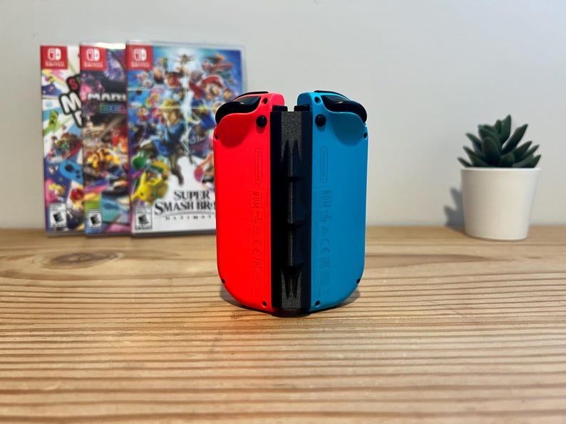 May include: A pair of red and blue Nintendo Switch Joy-Con controllers stand upright. Behind them are three game cases, with a small potted succulent plant to the right. The controllers are on a wooden surface.