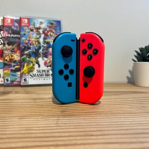 May include: A pair of blue and red Nintendo Switch Joy-Con controllers are displayed vertically. The controllers are in front of three game cases, with a small potted plant to the right. The game cases feature colourful graphics.