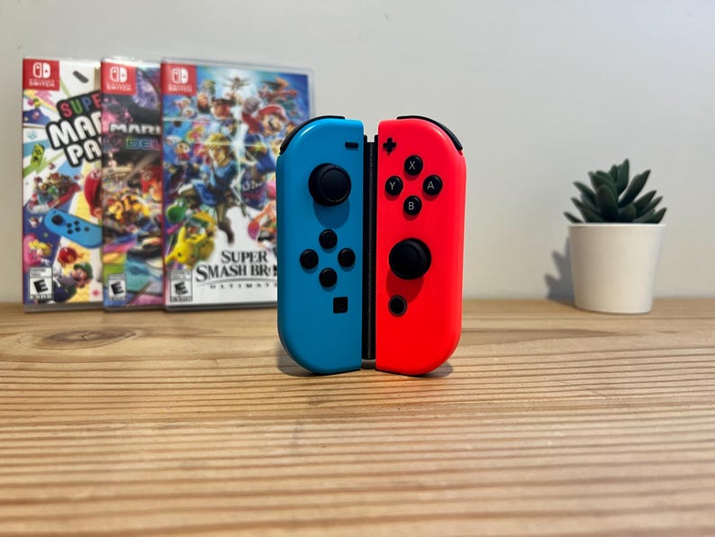 May include: A pair of blue and red Nintendo Switch Joy-Con controllers are displayed vertically. The controllers are in front of three game cases, with a small potted plant to the right. The game cases feature colourful graphics.