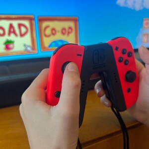 May include: A red and black Nintendo Switch controller is held in front of a television screen displaying a video game interface. The screen shows the words "LOAD" and "Co-op".