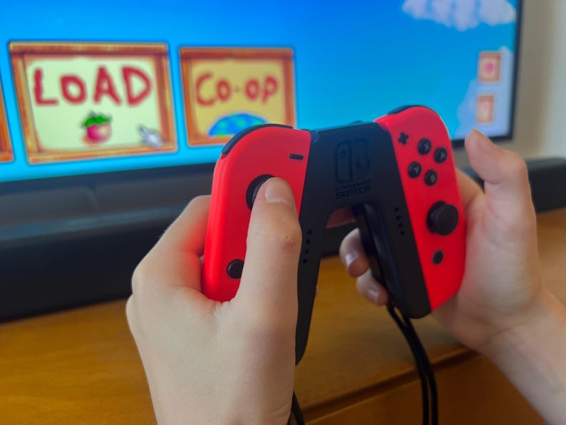 May include: A red and black Nintendo Switch controller is held in front of a television screen displaying a video game interface. The screen shows the words "LOAD" and "Co-op".