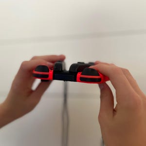 May include: A black and red video game controller with a sleek design. The controller has a black body with red accents on the sides and buttons. The controller is held in two hands.