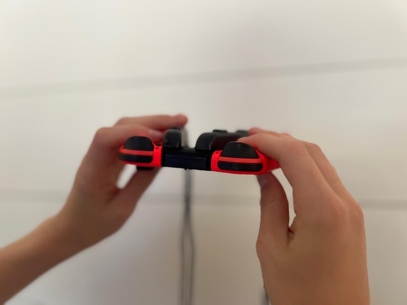 May include: A black and red video game controller with a sleek design. The controller has a black body with red accents on the sides and buttons. The controller is held in two hands.