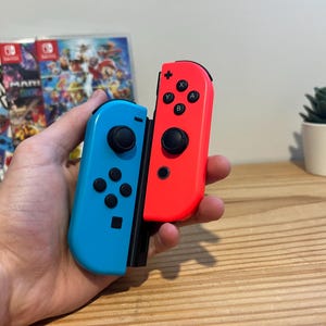 May include: A pair of Nintendo Switch Joy-Con controllers, one blue and one red, held in a hand. The controllers have black buttons and joysticks. A game case is visible in the background, along with a small potted plant.
