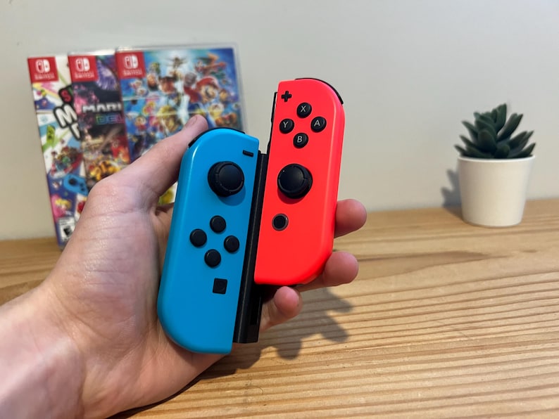 May include: A pair of Nintendo Switch Joy-Con controllers, one blue and one red, held in a hand. The controllers have black buttons and joysticks. A game case is visible in the background, along with a small potted plant.