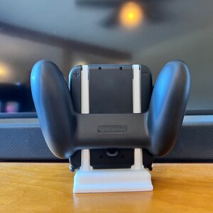 K&ouml;nnte beinhalten: Ein schwarzer Nintendo Switch Controller-Griff mit einem wei&szlig;en St&auml;nder. Der Controller ist an einer schwarzen Konsole befestigt. Der St&auml;nder ist wei&szlig; und rechteckig. Das Nintendo-Logo ist sichtbar.