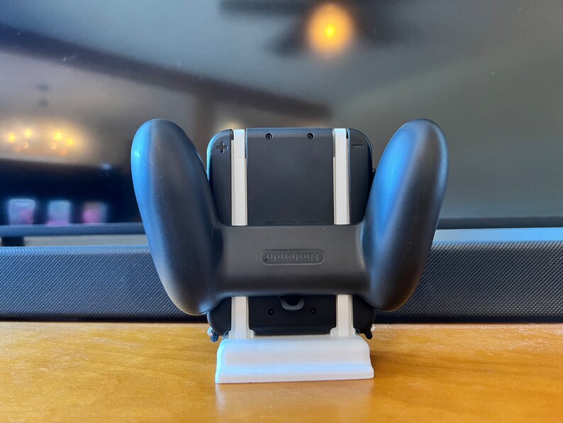 K&ouml;nnte beinhalten: Ein schwarzer Nintendo Switch Controller-Griff mit einem wei&szlig;en St&auml;nder. Der Controller ist an einer schwarzen Konsole befestigt. Der St&auml;nder ist wei&szlig; und rechteckig. Das Nintendo-Logo ist sichtbar.