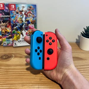 May include: A pair of blue and red Nintendo Switch Joy-Con controllers. The controllers are held in a hand, with game cases in the background. A small potted succulent is also visible. The controllers have black buttons and control sticks.