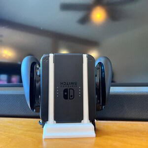 K&ouml;nnte beinhalten: Eine schwarze Nintendo Switch-Konsole mit angeschlossenen Joy-Con-Controllern, die in einem wei&szlig;en Ladest&auml;nder steht. Auf der Konsole steht das Wort "SWITCH". Die Controller sind schwarz und gebogen.