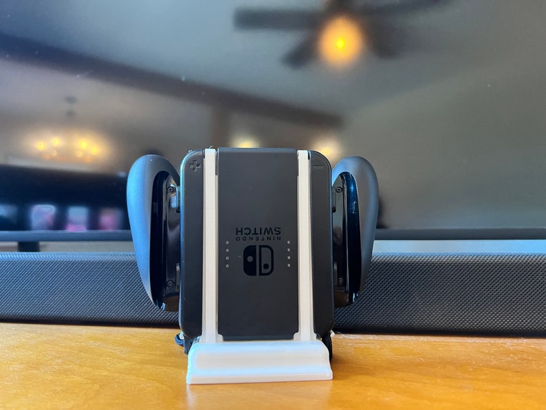 K&ouml;nnte beinhalten: Eine schwarze Nintendo Switch-Konsole mit angeschlossenen Joy-Con-Controllern, die in einem wei&szlig;en Ladest&auml;nder steht. Auf der Konsole steht das Wort "SWITCH". Die Controller sind schwarz und gebogen.