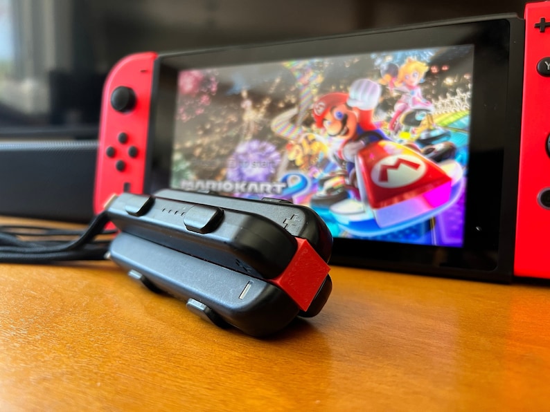 K&ouml;nnte beinhalten: Eine rote und schwarze Nintendo Switch-Konsole mit dem Spiel Mario Kart 8 Deluxe auf dem Bildschirm. Ein schwarzer Controller mit rotem Akzent im Vordergrund. Die Konsole steht auf einer Holzoberfl&auml;che.