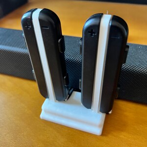 K&ouml;nnte beinhalten: Zwei schwarz-wei&szlig;e elektronische Ger&auml;te werden aufrecht in einem wei&szlig;en Halter angezeigt. Die Ger&auml;te haben einen wei&szlig;en Streifen in der Mitte und Plus- und Minuszeichen. Der Halter steht auf einer Holzoberfl&auml;che.