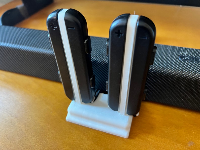 K&ouml;nnte beinhalten: Zwei schwarz-wei&szlig;e elektronische Ger&auml;te werden aufrecht in einem wei&szlig;en Halter angezeigt. Die Ger&auml;te haben einen wei&szlig;en Streifen in der Mitte und Plus- und Minuszeichen. Der Halter steht auf einer Holzoberfl&auml;che.