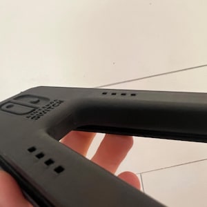 May include: A black Nintendo Switch accessory, possibly a controller grip or case, is held in a person's hand. The accessory has the Nintendo Switch logo and several small square vents. The item is made of a matte black material.