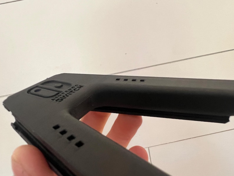 May include: A black Nintendo Switch accessory, possibly a controller grip or case, is held in a person's hand. The accessory has the Nintendo Switch logo and several small square vents. The item is made of a matte black material.
