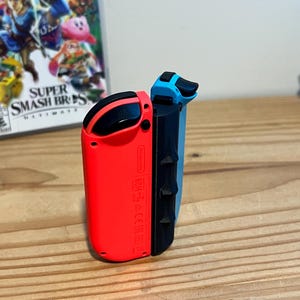 May include: A pair of Nintendo Switch Joy-Con controllers, one red and one blue, are shown. The controllers have black sides and buttons. The game case for "Super Smash Bros. Ultimate" is visible in the background.