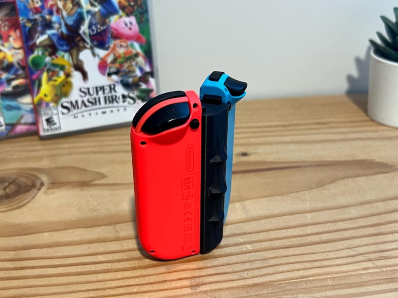 May include: A pair of Nintendo Switch Joy-Con controllers, one red and one blue, are shown. The controllers have black sides and buttons. The game case for "Super Smash Bros. Ultimate" is visible in the background.