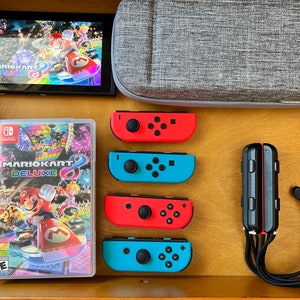 K&ouml;nnte beinhalten: Eine Nintendo Switch-Konsole mit dem Spiel "Mario Kart 8 Deluxe" auf dem Bildschirm und in einer Spielh&uuml;lle. Rote und blaue Joy-Con-Controller, eine graue Tragetasche und ein schwarzer Controller sind ebenfalls zu sehen.